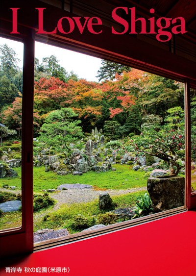 縦長の写真：赤い敷物が敷かれた和室の中から庭園が見える。庭園には苔むした岩と低木があり、地面は緑の草が生えている。岩の向こうには紅葉が色づいている。写真の上部に赤色の大きいアルファベットで「I Love Shiga」。左下に「青岸寺 秋の庭園（米原市）」