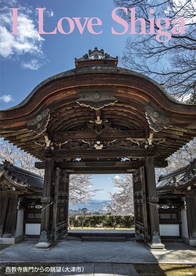 縦長の写真：青空をバックに真正面から撮影された古い立派な屋根付きの門がある。門の向こうには満開の桜と琵琶湖が見える。写真の上部にピンクの大きいアルファベットで「I Love Shiga」。左下に「西教寺唐門からの眺望（大津市）」