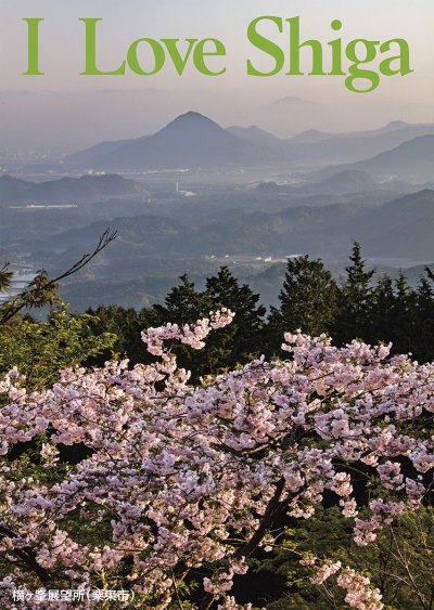 縦長の写真：下半分に満開の桜の枝のアップ。上半分は桜の向こうにある明け方のもやがかかった緑の山々を見下ろしている。写真の上部に黄緑色の大きいアルファベットで「I Love Shiga」。左下に「横ヶ峯展望所（栗東市）」