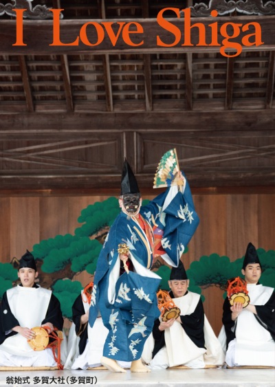縦長の写真：カキワリに松の木が描かれた舞台上の中央で、黒い翁の仮面をかぶり青い着物を着て烏帽子をかぶった男性が扇子を片手に舞っている。後ろには白い着物と烏帽子をかぶった男性が4人座っており、鼓を叩いている。写真の上部にオレンジの大きいアルファベットで「I Love Shiga」。左下に「翁始式 多賀大社（多賀町）」